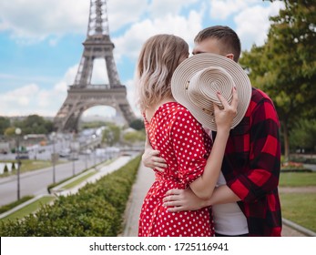 Young Beautiful Couple Kisses Covering Their Faces With A Straw Hat In Front Of Eiffel Tower In Paris. A Girl In A Red Dress With White Polka Dots And A Guy In A Red Plaid Shirt. Romantic Summer.