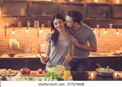 Young Beautiful Couple In Kitchen. Family Of Two Preparing Food. Man Kissing Woman While Making Delicious Fresh Salad. Nice Loft Interior With Light Bulbs