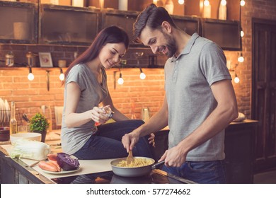 Young beautiful couple in kitchen. Family of two preparing food. Woman sitting on kitchen tabletop and helping man to make delicious pasta. Nice loft interior with light bulbs