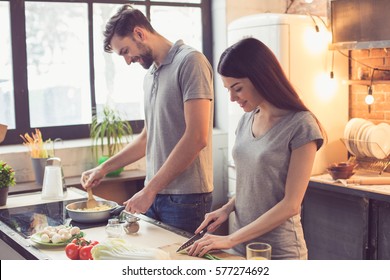 Young Beautiful Couple In Kitchen. Family Of Two Preparing Food. Couple Making Delicious Pasta. Nice Loft Interior With Light Bulbs And Big Window