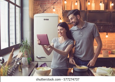 Young Beautiful Couple In Kitchen. Family Of Two Preparing Food. Couple Using Tablet Computer While Making Delicious Pasta. Nice Loft Interior With Light Bulbs And Big Window