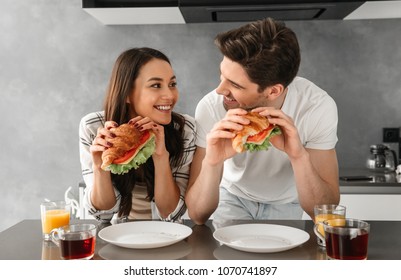 Young and beautiful couple looking at each other and smiling while eating sandwiches on breakfast in cozy house