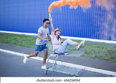 Young Beautiful Couple In Love, Boyfriend And Girlfriend Ride On Shopping Cart With Orange Smoke Bomb, Hipsters Having Fun In City Mall 