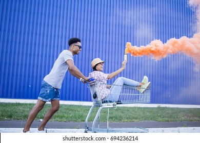 Young Beautiful Couple In Love, Boyfriend And Girlfriend Ride On Shopping Cart With Orange Smoke Bomb, Hipsters Having Fun In City Mall 