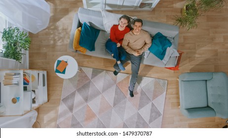 Young Beautiful Couple Is Lying Down On Sofa. Cozy Living Room With Modern Interior With Plants, Table And Wooden Floor. Top View.