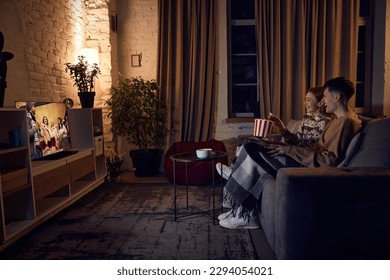 Young beautiful couple, man and woman spending time together at home in evening, sitting on couch, watching tv and eating popcorn. Concept of relationship, leisure activity, weekends, fun, emotions - Powered by Shutterstock