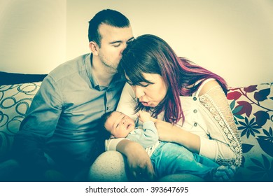 Young Beautiful Couple With New Baby At Home. Portrait Of A Smiling Happy Family With The Kid In Bright Domestic Background. Man And Woman With Newborn Child Sitting. Carefree Moment Of Happiness