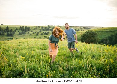 Young Beautiful Couple Red-haired Girl In A Pink Dress And Green Jacket A Man In A Gray T-shirt And Green Shorts Are Having Fun In The Grass In A Field In Nature At Sunset