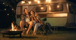 Young Beautiful Couple Relaxing At A Caravan Camping Area In The Evening, Keeping Warm With Campfire, Plaid Blanket And Warm Tea. Looking At The Stars And Wandering About A Successful Future Together.
