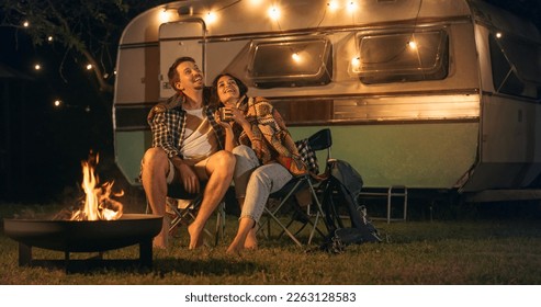 Young Beautiful Couple Relaxing at a Caravan Camping Area in the Evening, Keeping Warm with Campfire, Plaid Blanket and Warm Tea. Looking at the Stars and Wandering About a Successful Future Together.