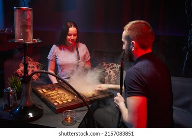 a young beautiful couple sits in a hookah lounge and plays backgammon, there is a hookah nearby, they smoke a hookah and exhale thick white smoke