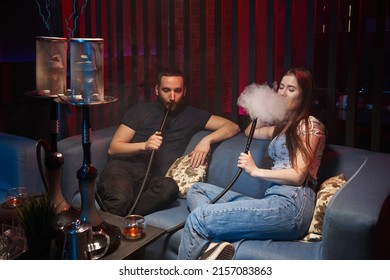 a young beautiful couple is sitting in a hookah lounge, there is a hookah nearby, they smoke a hookah and exhale thick white smoke