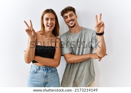 Young beautiful couple standing together over isolated background smiling with happy face winking at the camera doing victory sign with fingers. number two. 