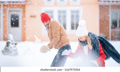 Young Beautiful Couple Throws Snowballs At Each Other While Snow Falls. Happy Man And Woman Playing With Snow In The Yard Of Their Idyllic House. Family Enjoying Winter.