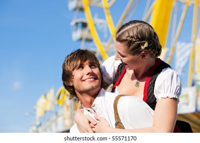 Young and beautiful couple in traditional Bavarian Tracht - Dirndl and Lederhosen - embracing each other on a fair like a Dult or the Oktoberfest; both are standing in front of a big Ferris wheel