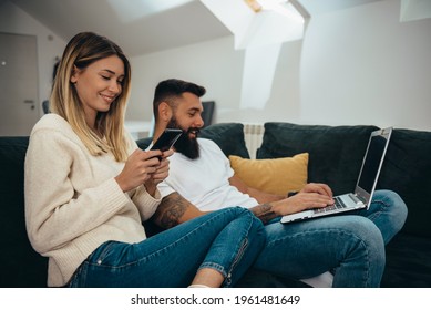 Young Beautiful Couple Using A Smartphone And A Laptop While Relaxing On The Couch At Home