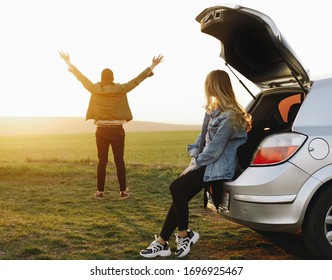 Young And Beautiful Couple, Who Look At And Show The Gesture Of Freedom At Sunset Near Their Car, With Open Trunk. Travel, Adventure, Lifestyle, Travel, Vacation Concept.