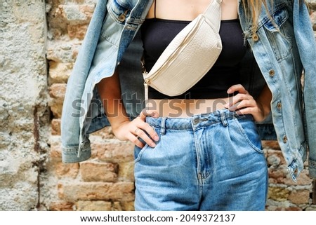 Young beautiful cropped woman standing next to street bricks wall, denim jeans clothes, holding belt satchel purse in shoulder, summer fashion trend, close-up details.