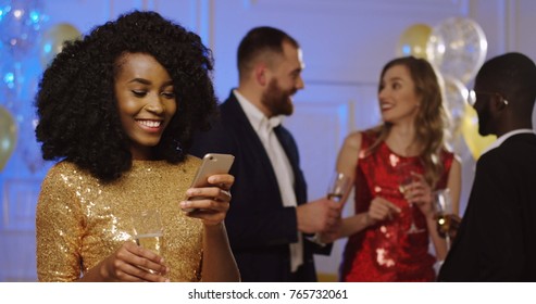 Young Beautiful Curly African American Woman In Glamorous Gold Dress Standing In The Middle Of The Party And Chatting On The Phone With Glass Of Champaigne In Her Hand. Dancing People And Balloons