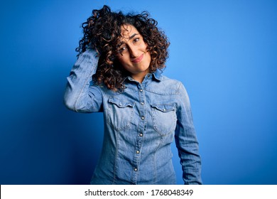 Young beautiful curly arab woman wearing casual denim shirt standing over blue background confuse and wonder about question. Uncertain with doubt, thinking with hand on head. Pensive concept. - Powered by Shutterstock