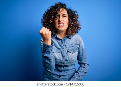 Young beautiful curly arab woman wearing casual denim shirt standing over blue background angry and mad raising fist frustrated and furious while shouting with anger. Rage and aggressive concept. - Shutterstock ID 1914715333