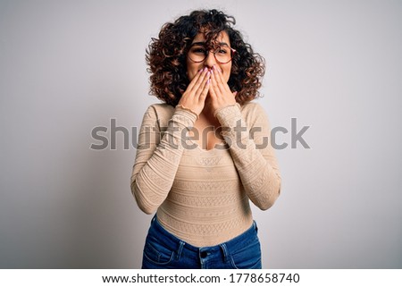 Young beautiful curly arab woman wearing casual t-shirt and glasses over white background laughing and embarrassed giggle covering mouth with hands, gossip and scandal concept