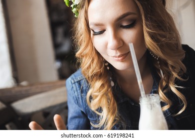 Young Beautiful Curly Blonde Bride With Bouquet Of Wreath Of Flowers On Head In White Wedding Dress In A Cafe With A Milkshake With A Straw In A Denim Jacket