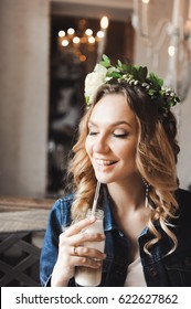 Young Beautiful Curly Blonde Bride With Bouquet Of Wreath Of Flowers On Head In White Wedding Dress In A Cafe With A Milkshake With A Straw In A Denim Jacket