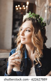 Young Beautiful Curly Blonde Bride With Bouquet Of Wreath Of Flowers On Head In White Wedding Dress In A Cafe With A Milkshake With A Straw In A Denim Jacket