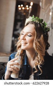 Young Beautiful Curly Blonde Bride With Bouquet Of Wreath Of Flowers On Head In White Wedding Dress In A Cafe With A Milkshake With A Straw In A Denim Jacket