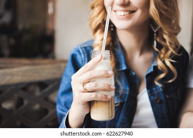 Young Beautiful Curly Blonde Bride With Bouquet Of Wreath Of Flowers On Head In White Wedding Dress In A Cafe With A Milkshake With A Straw In A Denim Jacket