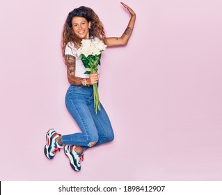 Young beautiful curly woman with tattoo holding bouquet of white rose smiling happy. Jumping with smile on face over isolated pink background