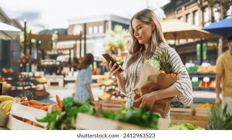 Young Beautiful Customer Shopping for Fresh Seasonal Fruits and Vegetables, Using Smartphone to Browse Internet on the Move. Female Holding a Sustainable Paper Bag with Ecological Local Farm Produce - Powered by Shutterstock