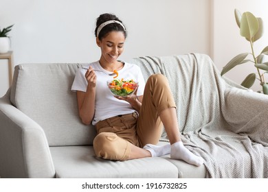 Young And Beautiful Dark-haired Lady Sitting On Sofa At Home, Eating Colorful Fresh Vegetable Salad And Smiling, Healthy Woman Enjoying Food Full Of Vitamins, Copy Space. Food, Diet, Healthy Lifestyle