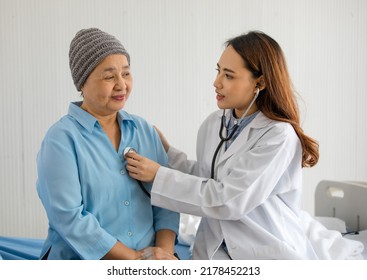 Young And Beautiful Doctor Using Stethoscope For Diagnosis With Older Asian Woman Patient Covered The Head With Clothes Effect From Chemo Treatment In Cancer Cure Process.
