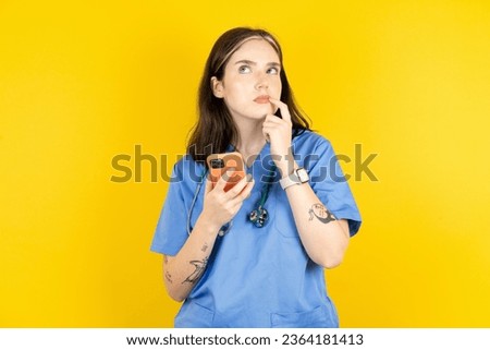 young beautiful doctor woman standing over yellow background thinks deeply about something, uses modern mobile phone, tries to made up good message, keeps index finger near lips.