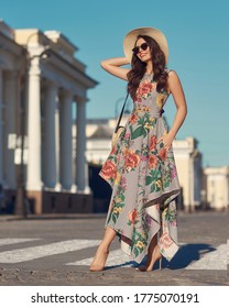 Young beautiful elegant tall slim woman with natural makeup and wavy brunete hair wearing colorful dress, salty hat and sunglassses walking in the city on a summer day and holding handbag