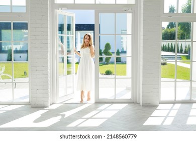 A Young Beautiful Elegant Woman Enters The Open Door Of A House With Huge High Panoramic Windows To The Floor, Through Which Rays Of Sunlight From The Street Will Come. Clean Bright White Room