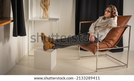Young, beautiful, emotional, surprised, business woman with glasses sitting on a chair in a bright room, tired from work, looking away. Places for inscriptions