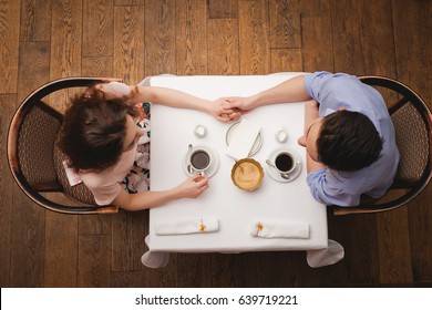 Young Beautiful Enamored Couple Sitting At A Table In A Cafe And And Holding Hands, View From Above