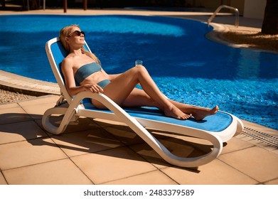 Young beautiful European girl sitting on a sun lounger by the pool, holding a glass with orange juice. Blonde girl relaxing in the fresh air by the pool. Concept of summer holiday and relaxation. - Powered by Shutterstock