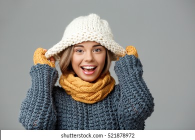 Young beautiful fair-haired girl in knited hat sweater scarf and mittens smiling looking at camera over grey background. 