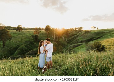 Young Beautiful Family With A Little Daughter And A Dog Hug, Kiss And Walk In Nature At Sunset.