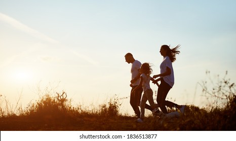 Young And Beautiful Family Of Three Are Jogging With Their Dog Outside The City On The Village Road On The Setting Sun, Side View, Copy Space