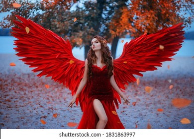 Young Beautiful Fantasy Woman Fallen Angel Stands Near Tree, Orange Falling Leaves.  Red Suit Costume Artificial Bird Wings Elegant Dress. Wind Magic Whirls Autumn Foliage. Girl Sexy Goddess Long Hair