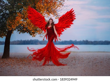 Young Beautiful Fantasy Woman Fallen Angel Lying In Air Near A Tree With Orange Leaves. Creative Red Costume, Huge Artificial Bird Wings And Elegant Dress. Magic Autumn Foliage. Photo Of Levitation