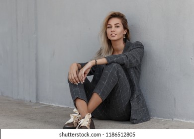 Young Beautiful Fashion Model Woman In A Gray Strict Vintage Suit With A Fashionable Jacket And Trousers Sits On The Street Near The Gray Wall