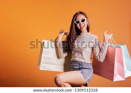 Young beautiful fashionable Asian woman holding shopping bags phone and credit card over orange background studio shot