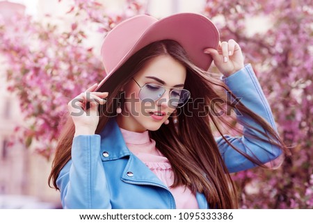 Young beautiful fashionable girl wearing stylish blue color aviator sunglasses, pink suede hat, earrings, biker jacket. Model posing in street with flowering trees. Spring fashion concept. 