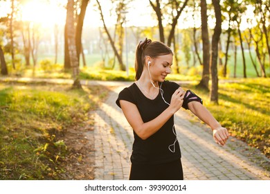 Young beautiful female athlete runner fixing her armband before training with sunset on background in park. Smiling girl with earbuds preparing for workout. Brightly lit photo with copy space - Powered by Shutterstock